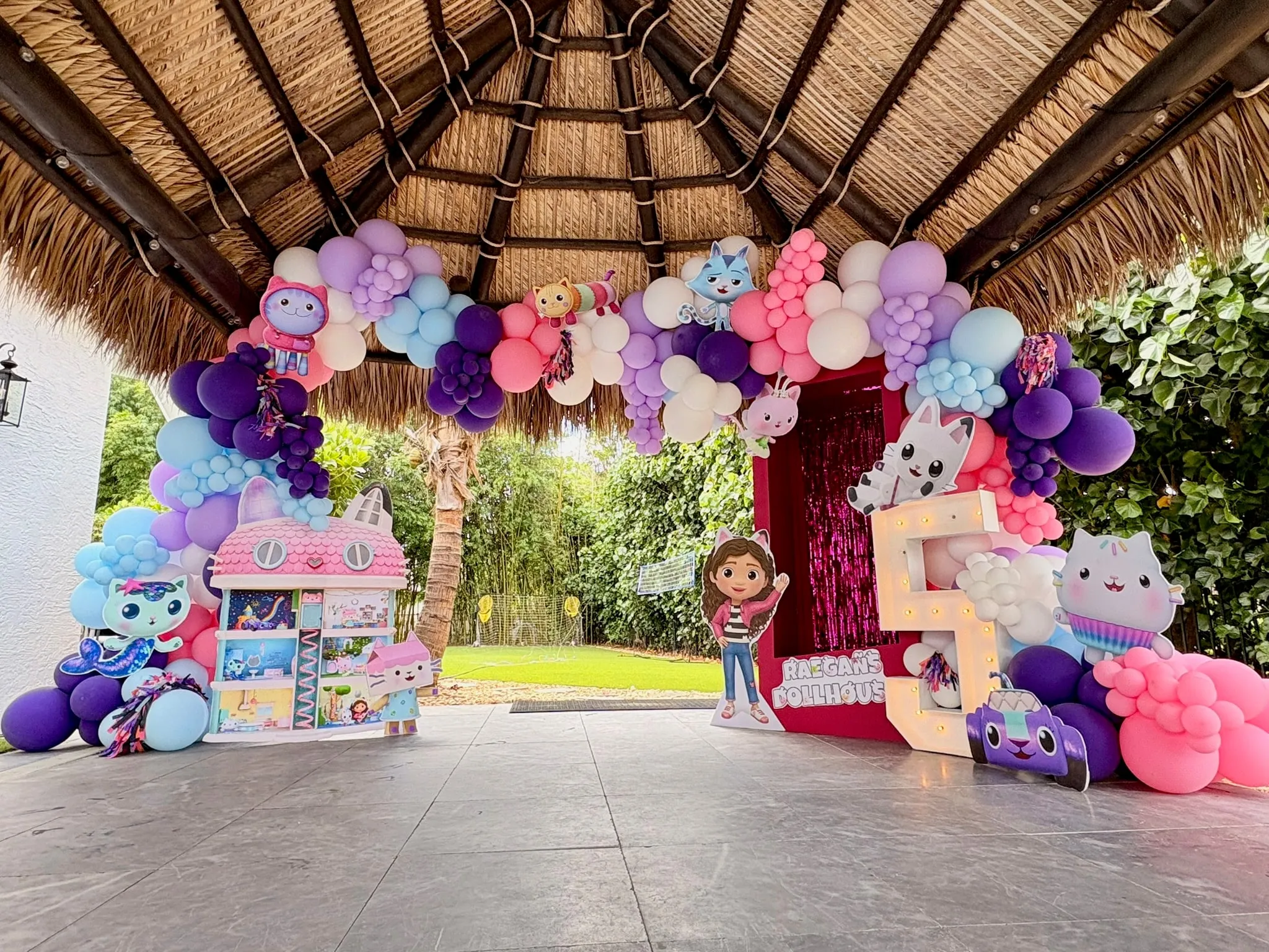 Pink and purple balloon arch under a thatched roof with a dollhouse, photo frame and light-up number five
