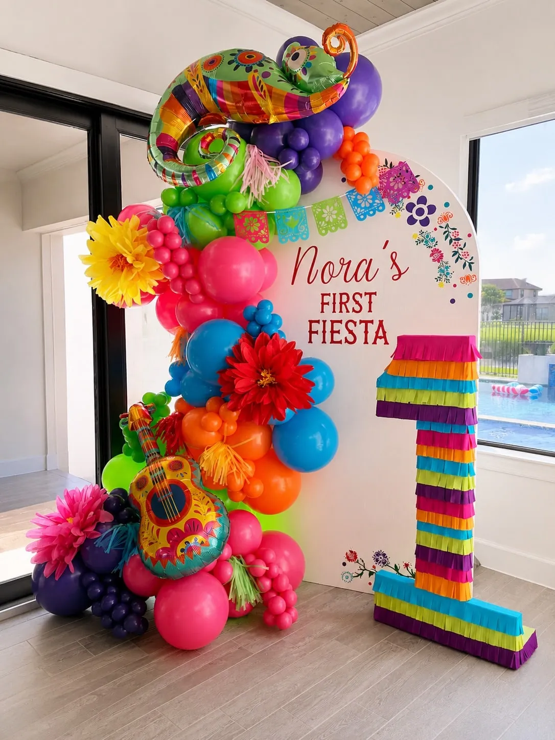 Bright multicolor balloon garland with paper flowers on an arched first birthday backdrop beside a fringed number one