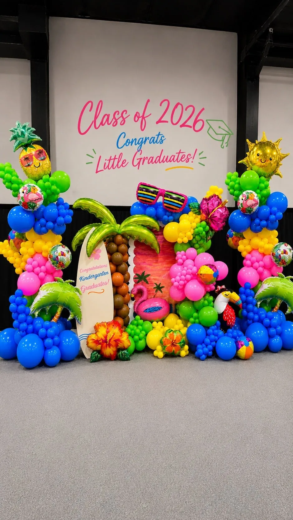 Stage decorated with bright balloon columns, palm tree balloons and a surfboard under a Class of 2026 screen