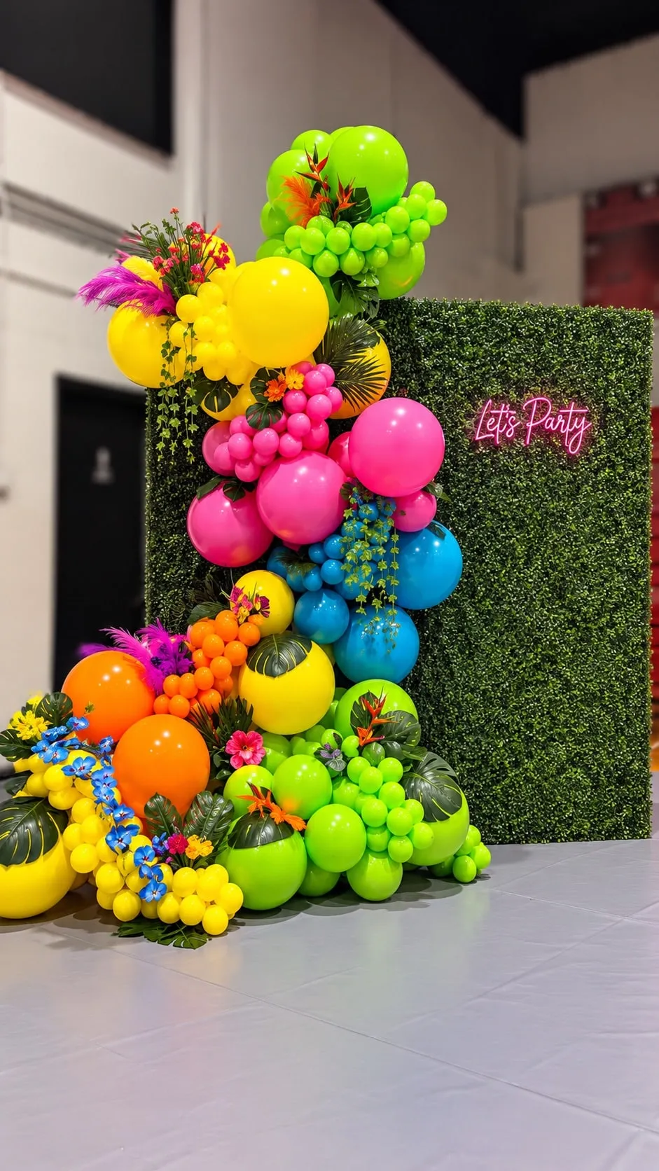 Neon pink, yellow, green and orange balloon garland with tropical flowers in front of a greenery wall