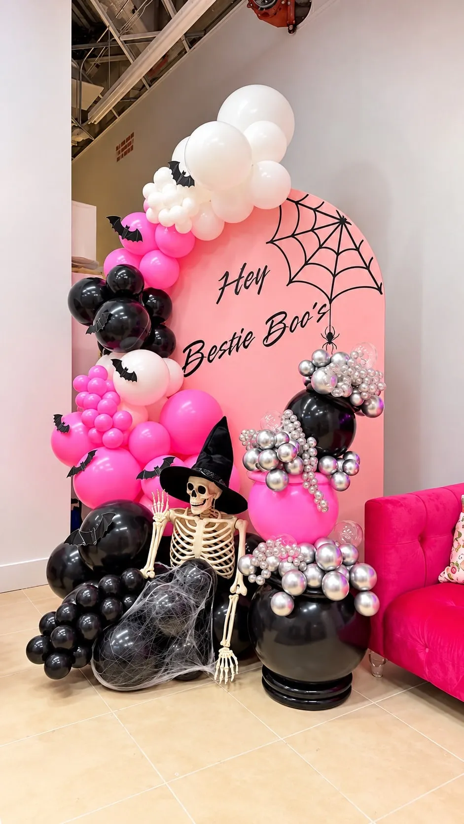 Pink arched backdrop with a spider web, framed by hot pink, black and white balloons with bats, silver bubble clusters and a skeleton in a witch hat
