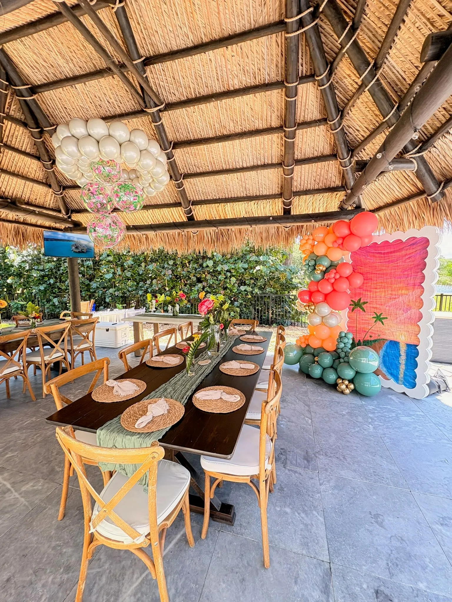 Dinner table under a thatched roof with a sunset backdrop, a coral and teal balloon garland and a ceiling balloon cluster